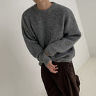Modell trägt einen grauen Pullover mit lockerer Passform und langen Ärmeln, kombiniert mit braunen Hosen.
